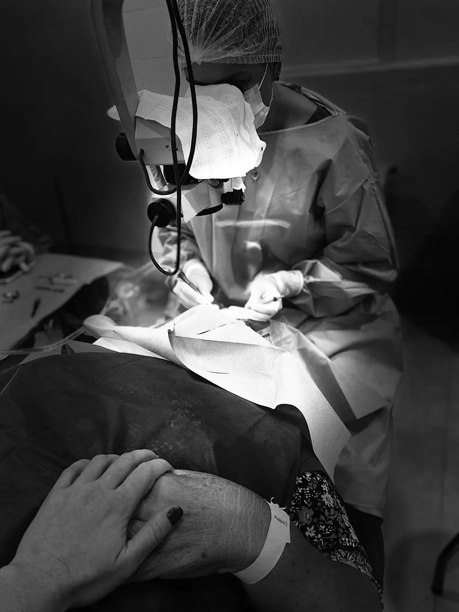 Durante a cirurgia de catarata, uma mão segura a mão do paciente enquanto a médica opera ao microscópio